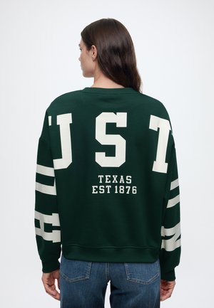 Person mit langen braunen Haaren, die einen dunkelgrünen Pullover mit weißen Buchstaben "UST" und "Texas Est 1876" auf dem Rücken trägt.