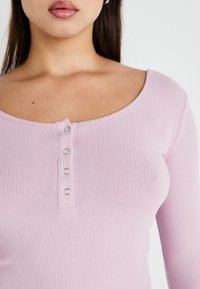 Lys rosa ribbet langermet topp med avrundet halslinje og fem sølvfargede trykknapper nedover foran, som fremhever et tettsittende design.