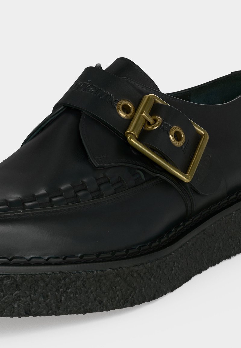 Schwarzer Lederschuh mit dicker strukturierter Sohle, dekorativen Nähten und großer Messingschnalle auf verstellbarem Riemen oben.