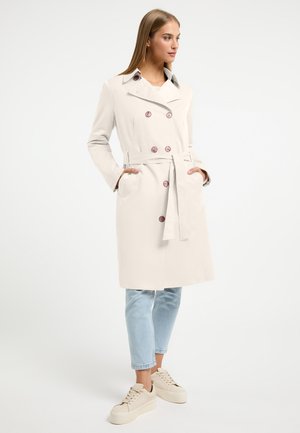 Beige doublé trapper met grote roze knopen, een ceintuur in de taille, zijzakken en een gekleurde kraag. Gecombineerd met lichtblauwe jeans en beige sneakers.