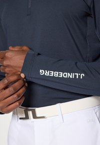 Granatowa koszula polo z długim rękawem i kołnierzykiem, z białym logo "J.Lindeberg" na mankiecie. W zestawie z białym paskiem i jasnymi spodniami.