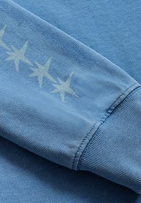 Jasnoniebieski rękaw swetra z teksturowanym materiałem i trzema graficznymi akcentami w kształcie gwiazdy w jaśniejszym odcieniu.