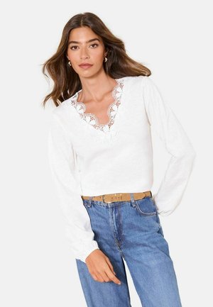 Woman with wavy brown hair wearing a white long-sleeve blouse with floral lace neckline and high-waisted blue jeans with tan belt.