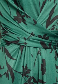 Grön blommig tyg med svarta accenter, som har en rynkad design och slät textur, framhäver klänningens drapering och silhuett.