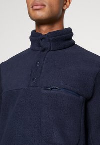 Sudadera de forro polar azul marino con cuello alto, botones a presión y un bolsillo en el pecho con cremallera. Presenta una textura suave y un logo de la marca discreto.