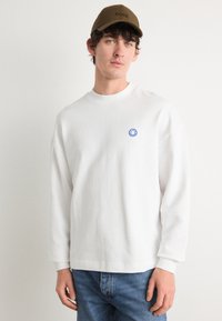 Camisa blanca de manga larga con cuello redondo, con un pequeño logo de cara sonriente azul en el pecho. Combinada con unos vaqueros azules y una gorra marrón.