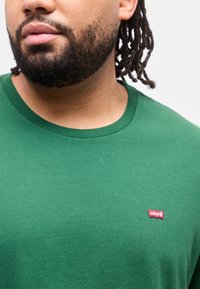 Άνδρας με γενειάδα και dreadlocks φορώντας πράσινο t-shirt Levi's, που δείχνει το κόκκινο λογότυπο Levi's στο στήθος.