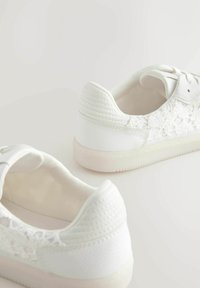 Sneakers bianche con pannello laterale in pizzo strutturato e suola in gomma liscia. Il tallone è leggermente imbottito con un design cucito.