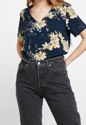 Chemise bleu marine à col en V avec imprimé floral jaune rentrée dans un jean noir taille haute sur une personne les mains derrière le dos.