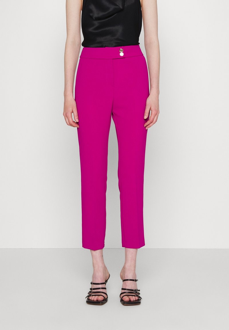 Vrouw in helderroze cropped broek, zwart mouwloos topje en zwarte stiletto sandalen, staand voor een effen achtergrond.