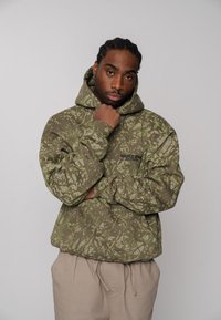 Sweat à capuche vert olive avec des motifs abstraits vert clair, doté d'une poche kangourou et d'une capuche à cordon ; associé à un pantalon beige.