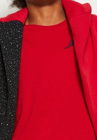 Červené tričko s krátkým rukávem, s černým vyšitým logem. Vedle je černá bunda s tečkovaným vzorem v světle modré barvě.