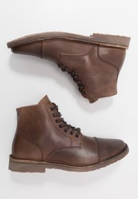 Hnědé kožené kotníkové boty s kulatou špičkou, šněrovacím designem a kontrastní texturovanou špičkou a patou. Pevná podrážka s protiskluzovým vzorem.
