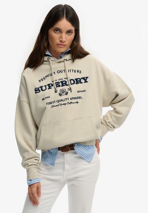 Kobieta w beżowej bluzie Superdry na suwak, nałożonej na niebieską koszulę w paski, oraz białe spodnie, stojąca z jedną ręką na biodrze na białym tle.