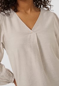 Blouse beige clair avec encolure en V, fronces aux épaules et longues manches bouffantes. Tissu lisse avec une texture subtile.