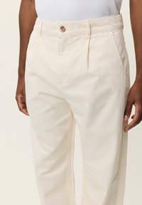 Φωτεινό μπεζ παντελόνι ραμμένο στο μέτρο με πιέτα μπροστά, κλείσιμο με κουμπί και πλευρικές τσέπες, κατασκευασμένο από απαλό βαμβακερό ύφασμα.