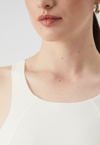 Bijela bezrukavna majica glatke teksture, s okruglim izrezom i suptilnim detaljima šava. Tono kože vidljivo oko područja vrata.