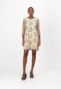 Abito a maniche corte con motivo floreale in oro e nero su sfondo beige, caratterizzato da un colletto rotondo e un orlo orlato. Scarpe con tacco a blocco nere.