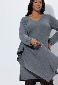 Vestido gris de punto con escote en V y diseño acampanado, con mangas largas y detalles de tela fluida alrededor del dobladillo.