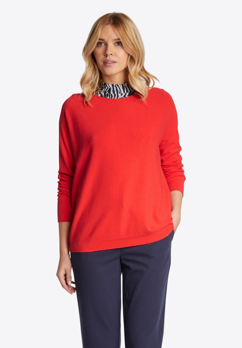 Roter Strickpullover mit rundem Ausschnitt und langen Ärmeln, über einem schwarz-weißen gestreiften Rollkragenpullover getragen. Glatte Textur, lässige Passform.