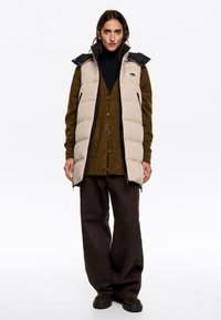 Gilet imbottito beige con colletto alto e dettagli neri, indossato sopra un maglione verde scuro e pantaloni larghi marroni. Presenta tasche con zip.