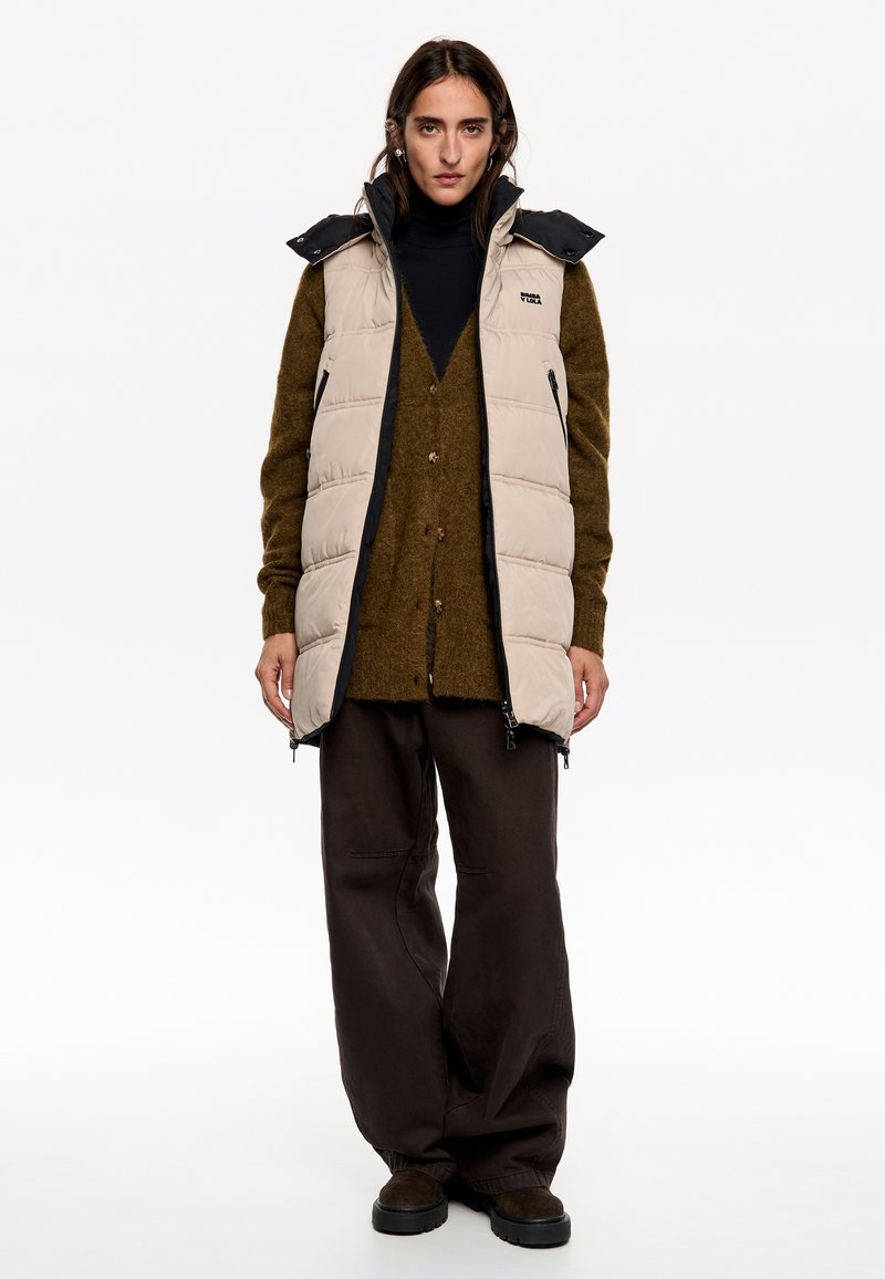 Gilet imbottito beige con colletto alto e dettagli neri, indossato sopra un maglione verde scuro e pantaloni larghi marroni. Presenta tasche con zip.