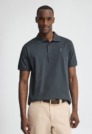 Hombre que lleva una camisa polo de manga corta gris oscuro y pantalones beige, de pie frente a un fondo liso y claro, mirando hacia adelante.