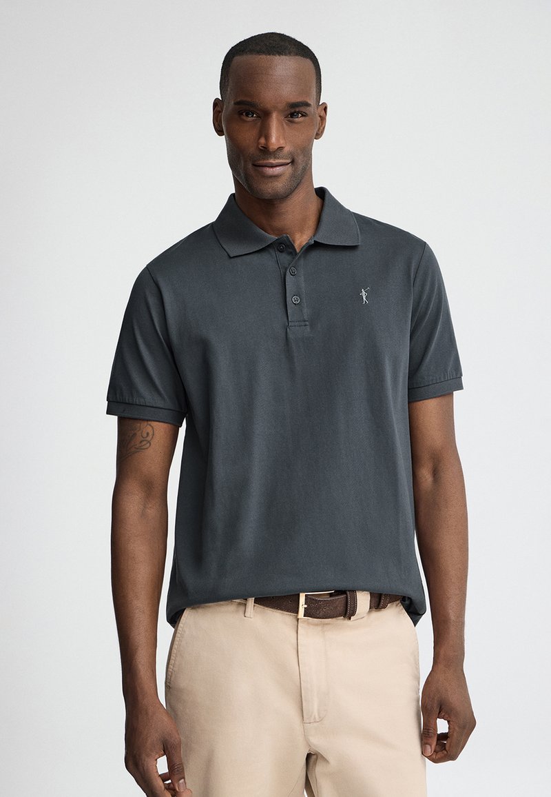 Homme portant un polo gris foncé à manches courtes et un pantalon beige, debout devant un fond clair uni, faisant face à l'avant.