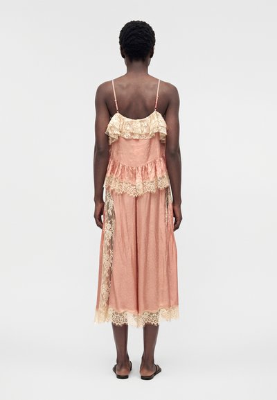 Γυναίκα με κοντά σγουρά μαλλιά φοράει ένα ροζ σατέν μίντι φόρεμα χωρίς μανίκια με μπεζ δαντελωτές βολάν και μαύρα φλατ σανδάλια, να στέκεται με πλάτη.