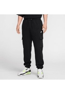 Mężczyzna ubrany w czarne spodnie dresowe cargo z logo Nike i białe buty sportowe z neonowo zielonymi akcentami, stojący na jednolitym tle.