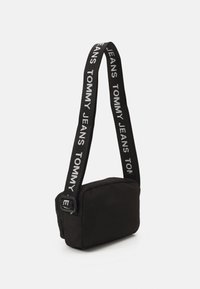 Černá obdélníková crossbody taška s nastavitelným popruhem, na kterém se opakuje bílé textové logo "TOMMY JEANS".