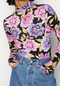 Langärmliges Oberteil mit Stehkragen in Schwarz, mit großem pinken und blauen Blumenmuster. Kombiniert mit hochgeschnittenen Jeans-Shorts mit Knopfverschluss.
