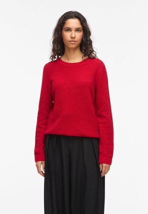 Kvinde med mellemtonet hud og krøllet mørkt hår iført en tekstureret rød sweater og brede sorte bukser, stående mod en ensfarvet hvid baggrund.