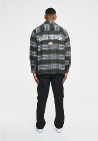 Green plaid flannel jacket with long sleeves, featuring a white embroidered logo and text on the back. Paired with black cargo pants.