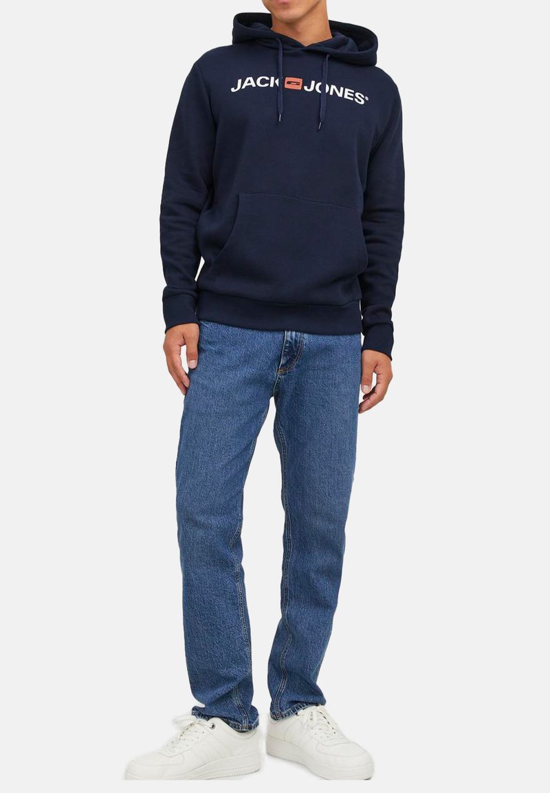 Mann trägt eine dunkelblaue Kapuzenjacke mit dem "JACK & JONES"-Logo, blaue Jeans und weiße Turnschuhe und steht vor einem einfarbigen Hintergrund.