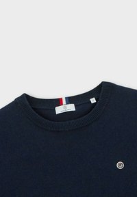 Pull en maille bleu marine avec un col rond côtelé et un petit écusson circulaire sur la poitrine. L'étiquette indique "Serge Blanco", taille M.