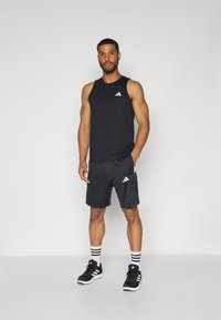 Black athletic tank top with a white logo, paired with black shorts featuring a side pocket and white stripes. Black sneakers complete the outfit.