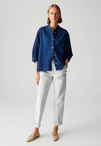 Chemise en denim bleu foncé avec un col mandarin, fermeture à boutons et coupe ample, associée à un pantalon blanc retroussé et des ballerines beiges.