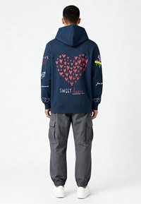 Sweat à capuche bleu marine avec un grand graphisme de cœur rouge au dos, accompagné d'un texte manuscrit "SWEET heart" et de divers accents colorés. Un pantalon cargo gris complète la tenue.