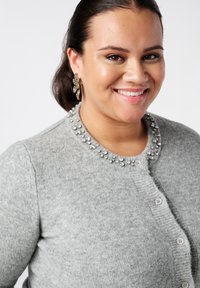 Grijze cardigan gemaakt van zacht materiaal met een ronde hals versierd met zilveren versieringen en een knoopsluiting aan de voorkant.