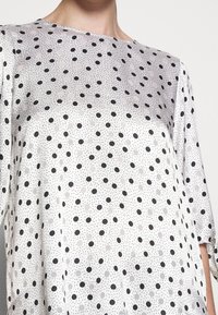 Witte satijnen blouse met zwarte en lichtgrijze stippen, met een ronde hals en driekwartmouwen met strikjes.