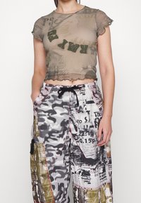 Przezroczysty oliwkowy top na krótko z falbankami, zestawiony z wzorzystymi szerokimi spodniami w kolorze czarnym, białym i ziemistych odcieniach, z wieloma grafikami.
