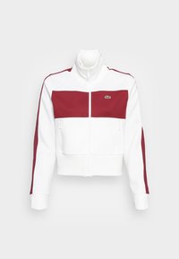 Vit och vinröd zip-up träningsjacka med hög krage, dragkedjefickor på sidan och en liten broderad grön krokodil-logotyp på bröstet.