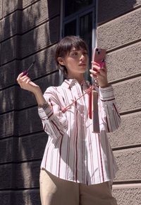Camisa a rayas blancas y rojas, pantalones beige, sosteniendo una máscara de pestañas morada y un teléfono rosa. También incluye un peine con un borla roja adjunta.