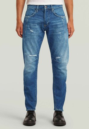 Homme portant un jean bleu délavé avec de petites déchirures aux cuisses et aux genoux, une chemise blanche rentrée et des bottes noires à lacets, debout sur un fond clair.