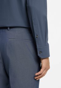 Man draagt een marineblauw overhemd en bijpassende op maat gemaakte broek, hand ontspannen langs het lichaam, tegen een effen witte achtergrond.