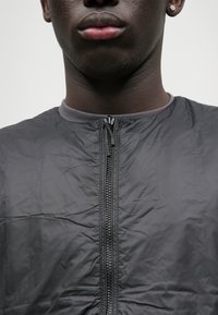 Γκρίζα ζακέτα με φερμουάρ και στρογγυλή λαιμόκοψη, κατασκευασμένη από υφασμάτινη, ελαφριά ύλη και με ορατό φερμουάρ μπροστά.