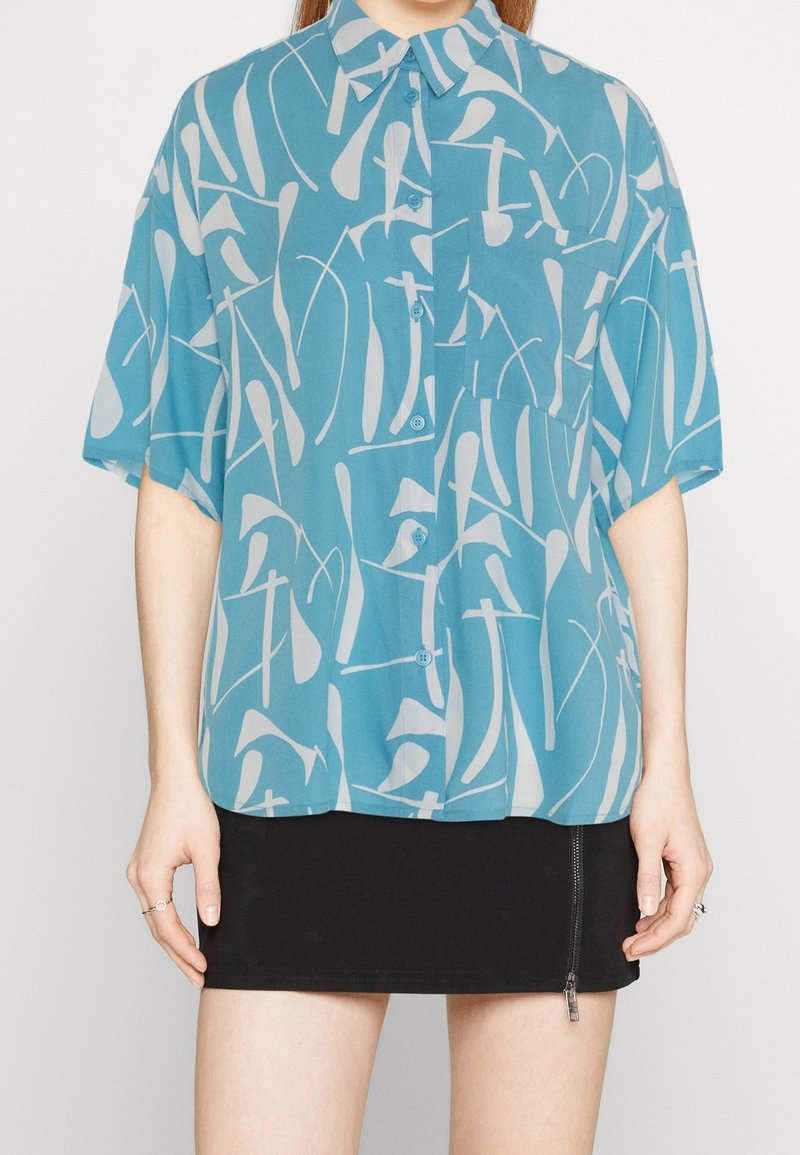 Una camicia a maniche corte, con bottoni, di colore azzurro chiaro con un motivo astratto bianco; realizzata in tessuto leggero; presenta una tasca singola.
