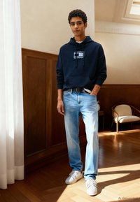 Noor mees seisab toas, kandes tumesinist kapuutsi, helesiniseid teksapükse ja valgeid tenniseid, ühe käega taskus päikesepaistelises toas.