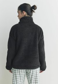 Donkerbruin, donzige fleece pullover met een hoge nek en een relaxte pasvorm, gedragen met witte geruite broeken. Zachte textuur en losse mouwen.